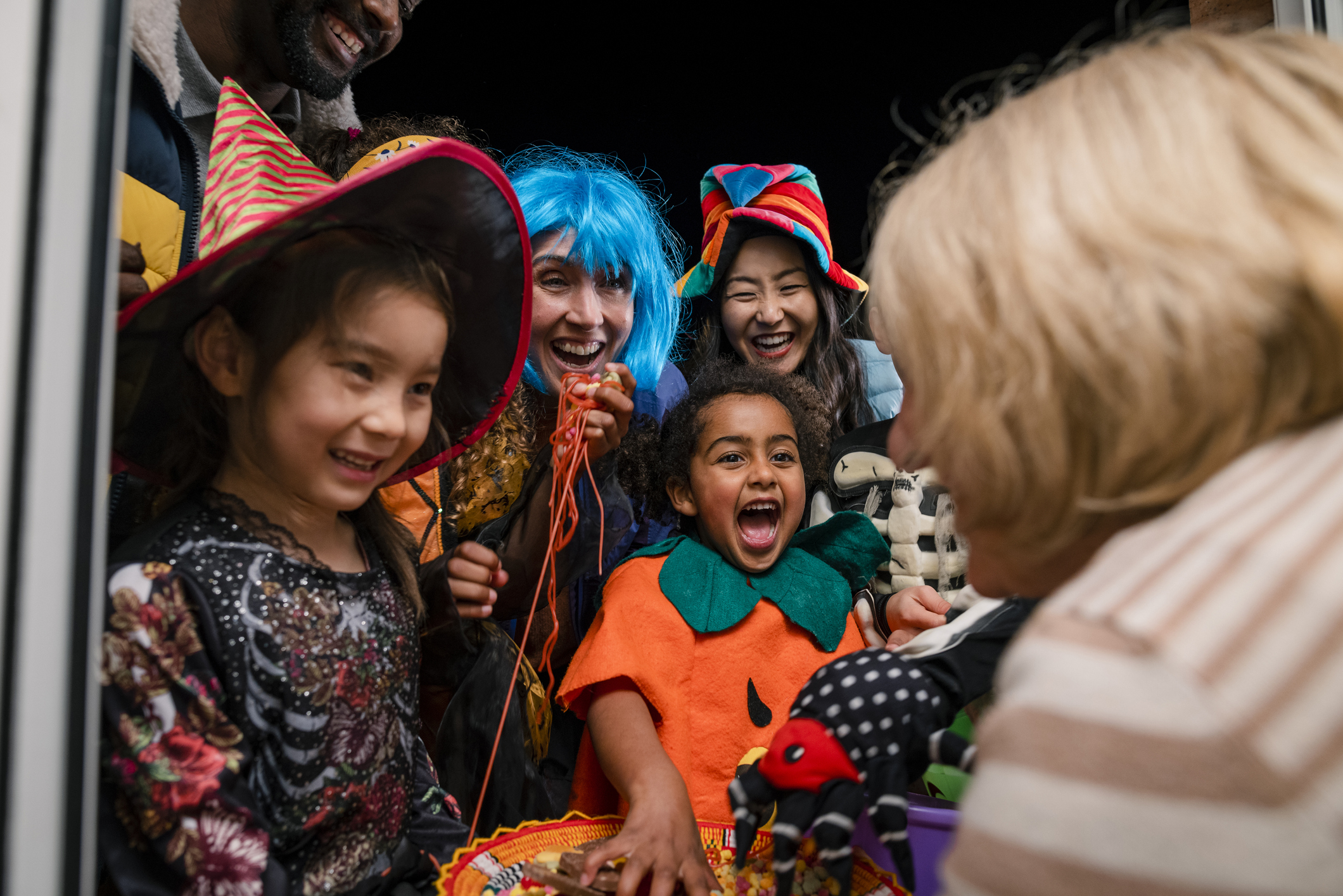 Taken from inside a residential house, family of trick-or-treaters treating in on halloween. They are taking sweets off a plate that an senior woman is holding.