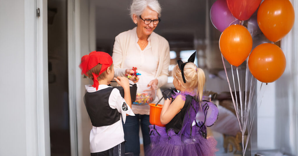 senior woman handing out candy to two children on Halloween dressed up in costumes.