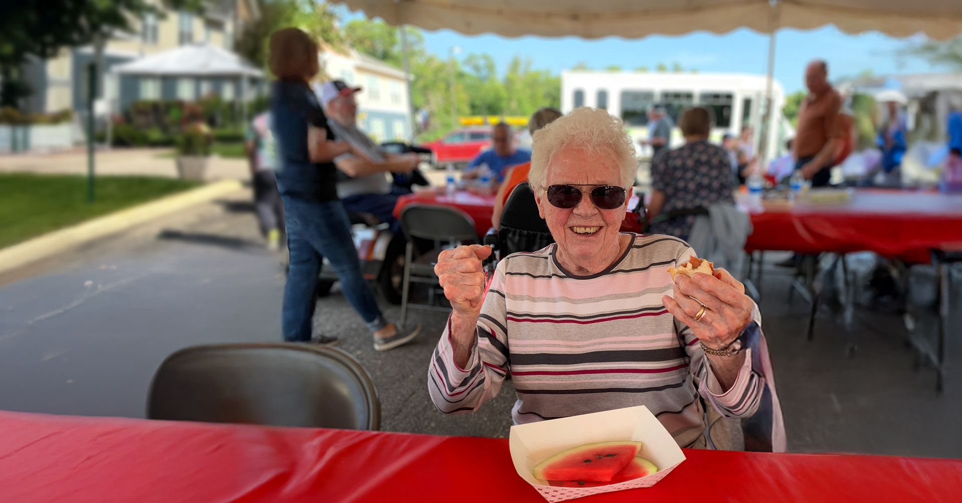 senior woman sitting at a folding table outside under a large event tent at a summer celebration enjoying a hot dog and watermelon