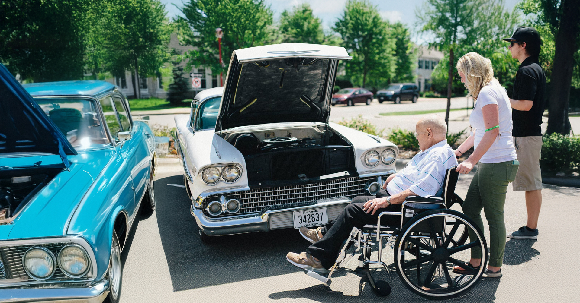 a senior man in a wheelchair being wheeled through a parking lot by a woman and a man outside on a sunny day attending a classic car show