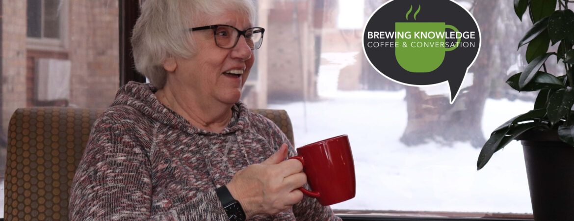 Elderly person with glasses smiles while holding a red mug indoors. Snowy view outside. Logo beside them reads "Brewing Knowledge Coffee and Conversation."