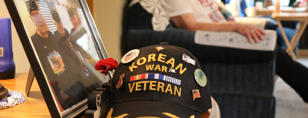A black Korean War veteran cap with pins sits on a wooden table next to a framed photo of veterans,