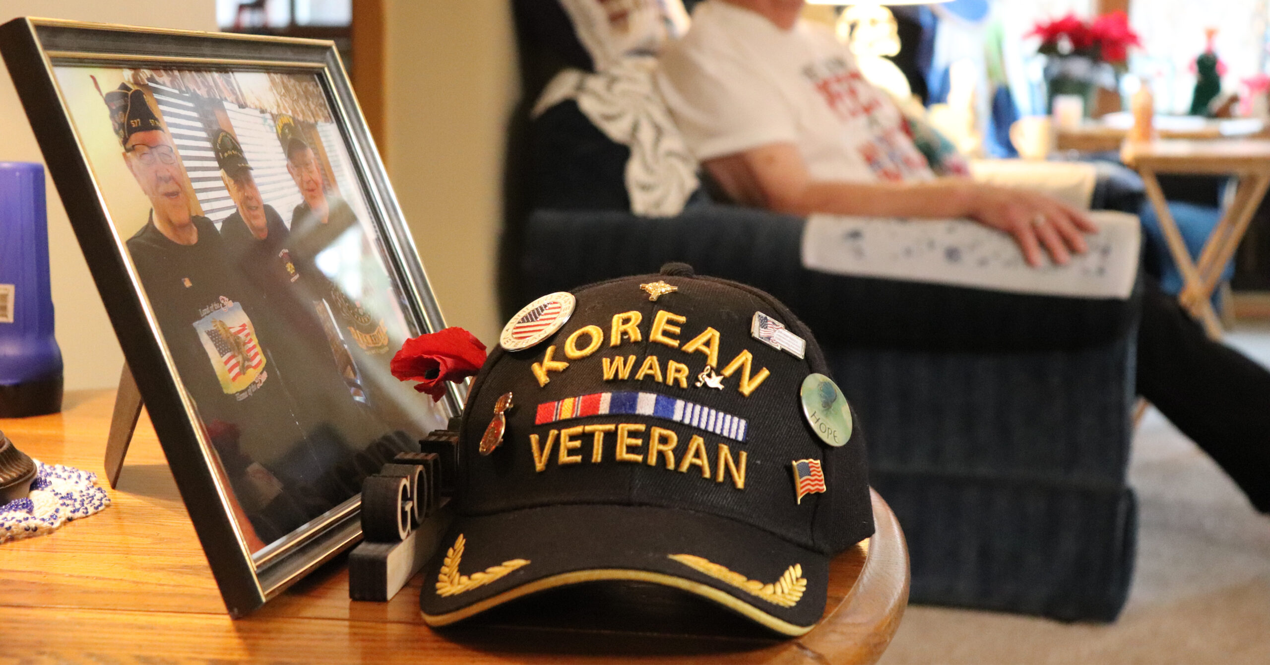 A black Korean War veteran cap with pins sits on a wooden table next to a framed photo of veterans,